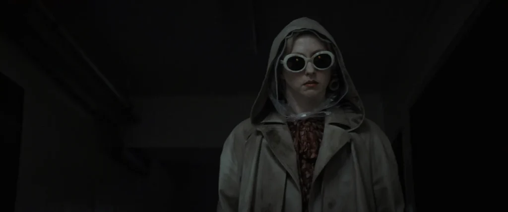 Image tirée du film Cuckoo, on y voit une dame en grand manteau trensh, la tête encapuchée, des lunettes noire, dans une pièce sombre.