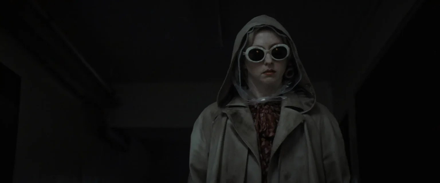Image tirée du film Cuckoo, on y voit une dame en grand manteau trensh, la tête encapuchée, des lunettes noire, dans une pièce sombre.