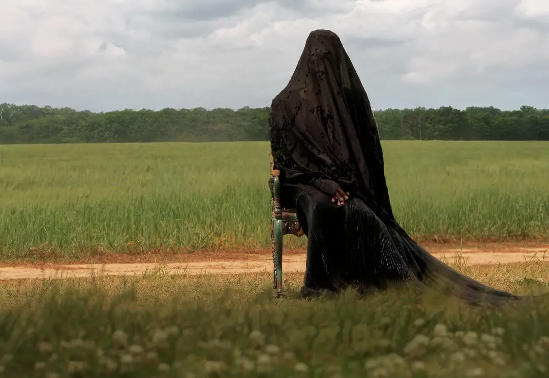 Une femme assise sur un siège dans le jardin d'une propriété privée, il fait jour et elle est entièrement recouverte par un voile noir.
