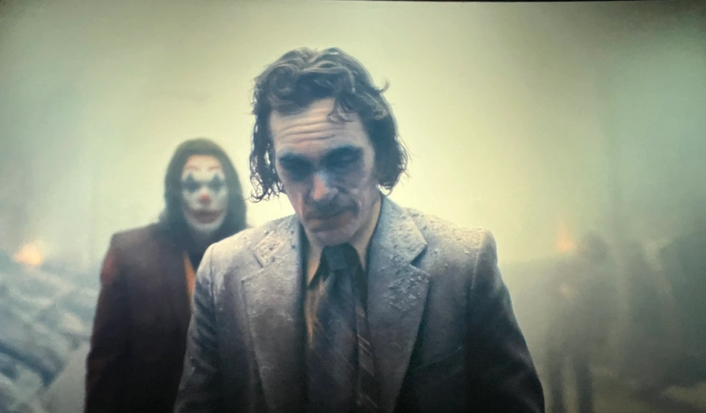 Joaquin Pheonix en joker marchant vers la caméra dans u nuage de poussière jaune, un autre joker le regarde par derrière.