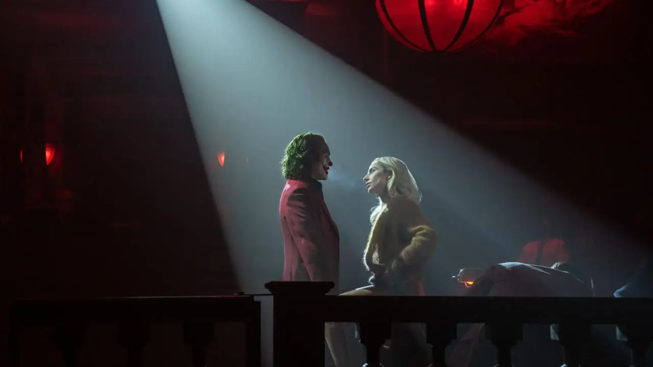 Lady Gaga en Harley Quinn et Joaquin Phoenix en Joker sur une scène, dans un halo de lumière avec, un garde fou dans l'ombre du premier plan laisse pensé qu'il sont dans un genre de tribunal ayant des airs de théâtre music hall.
