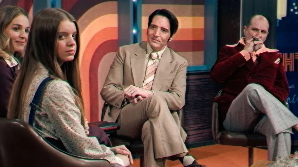 Un présentateur TV, regarde ses invités sur son plateau style années 70s.