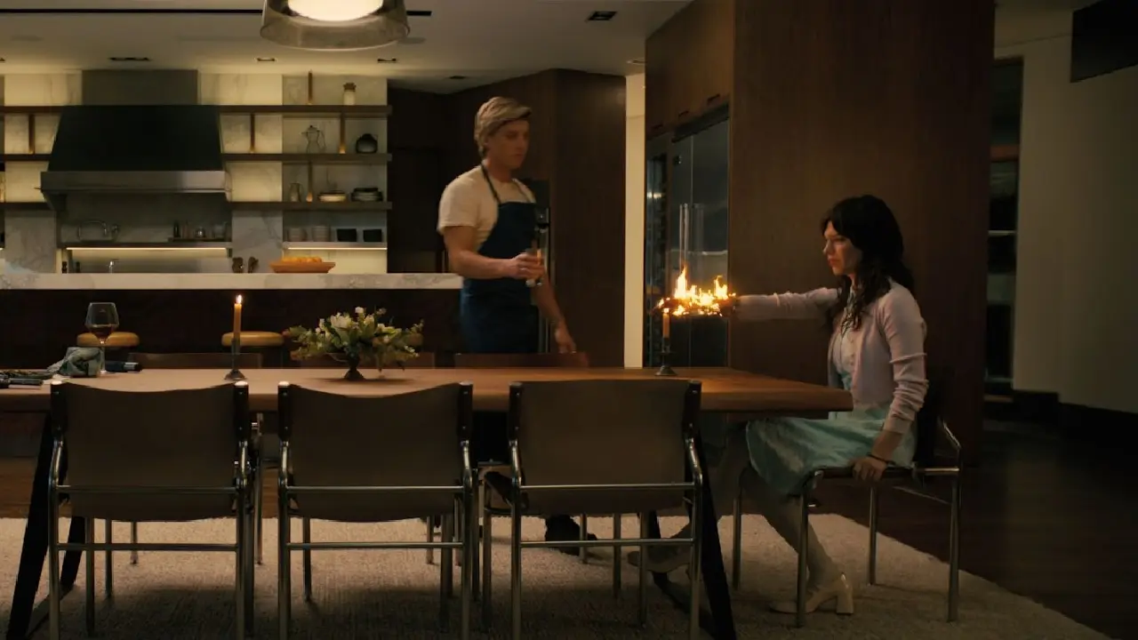 Image tirée du film Companion, on y voit Iris, une femme robot, assise de profil devant une table, elle tend la main vers une bougie et prend feu, un homme vient de la cuisine en arrière blanc avec un verre d'eau, très sereinement.