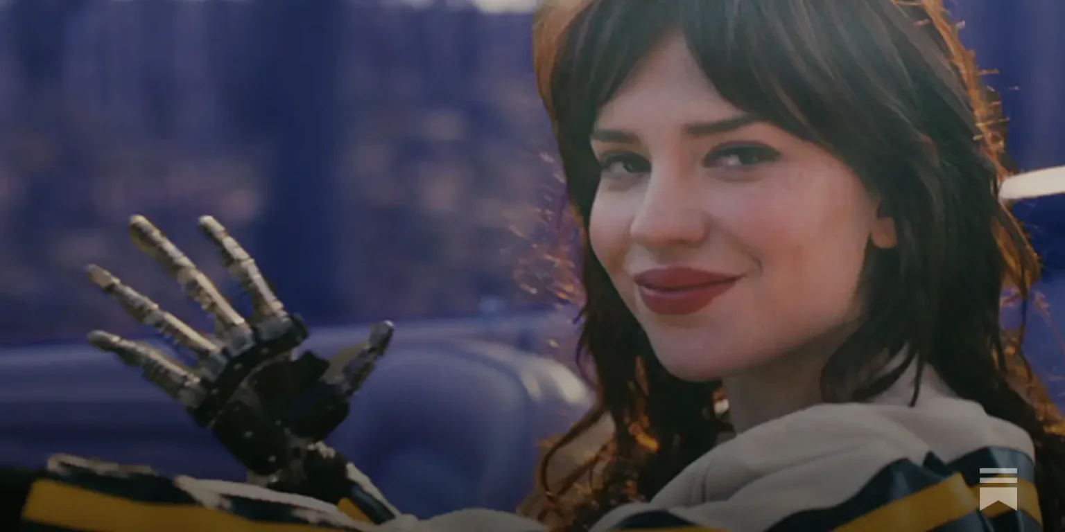 Image tire du film Companion, on y voit une femme en gros plan dans une voiture décapotable sur la route, saluer la caméra avec une main robotique.