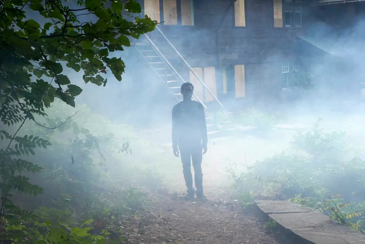 Image tirée du film It comes At night, on y vot un jeune homme devant une maison en pleine forêt, la nuit et dans le brouillard.