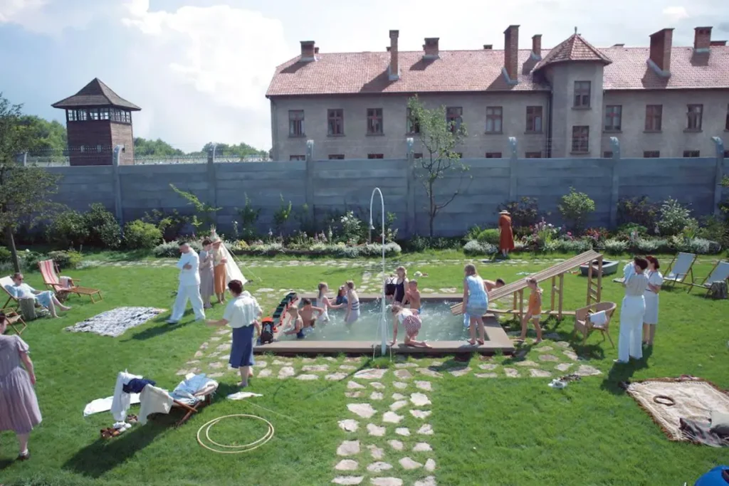 Des enfants jouent dans une piscine au beau milieu d'un magnifique jardin ensoleillée, au fond, l'horreur des camps de concentration d'Auschwitz.