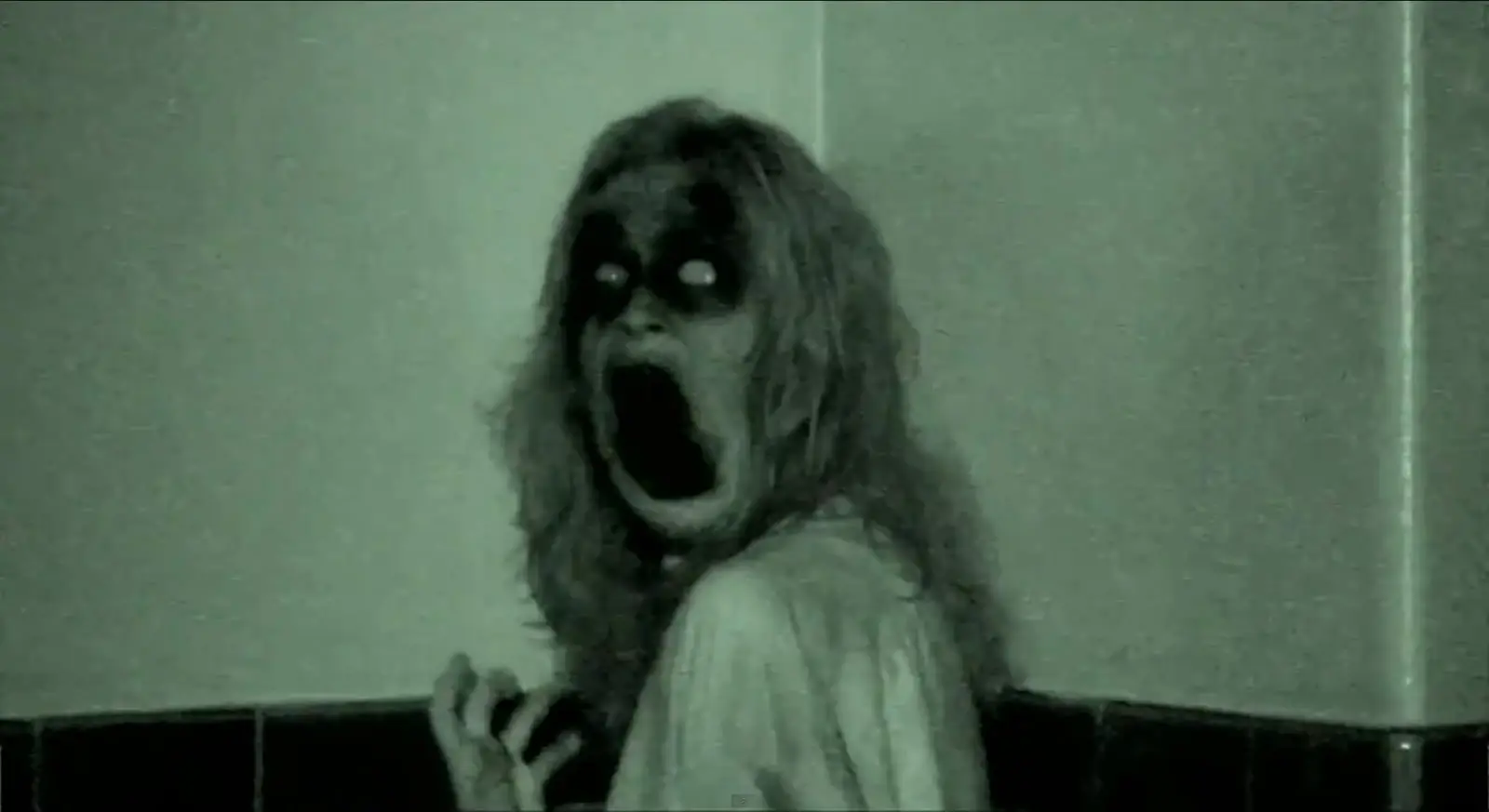 Une femme dans ce qui semble être un hôpital abandonné se retourne, elle a le visage noir et la bouche déformée. Le grain est vert et de mauvaise qualité.