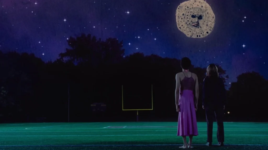 Deux jeunes femmes son devant un terrain de basket, la nuit, et regarde en direction de la lune qui possède un visage.