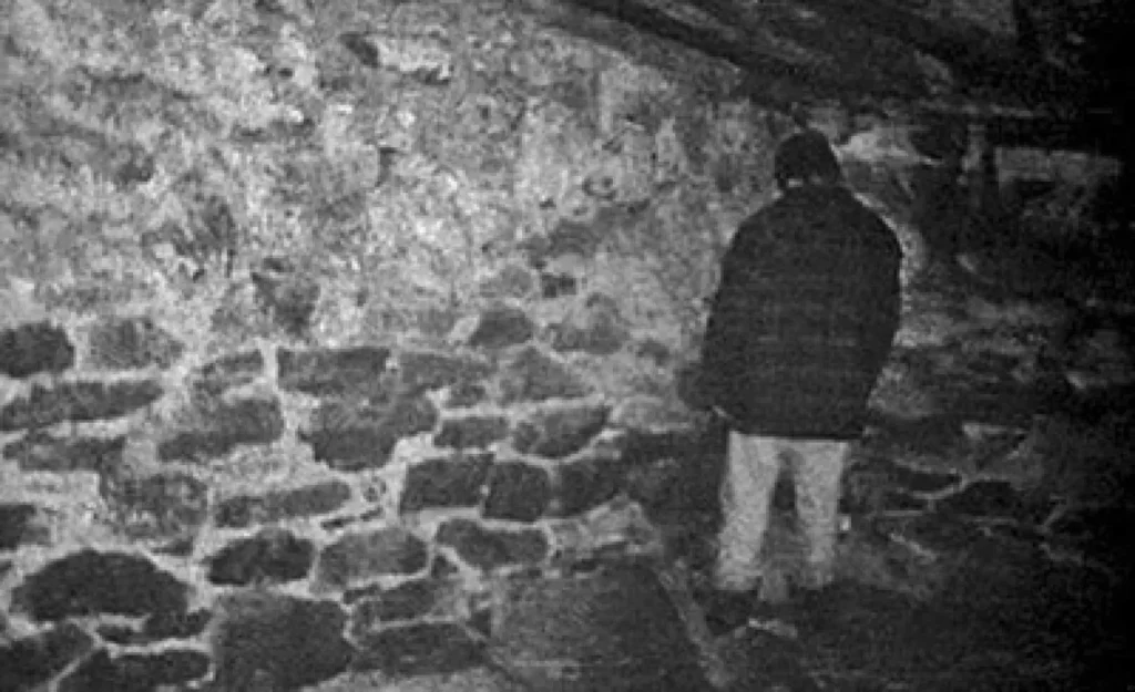 Image faisant pensé à l'esthétique cursed. Un homme est debout face à un mur dans une grange.