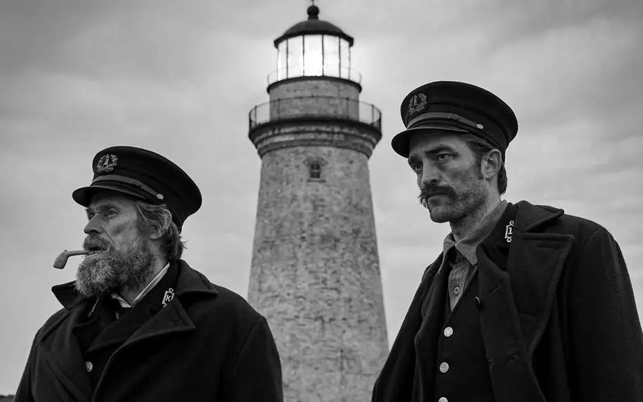 Photographie en noir et blanc, deux hommes, l'un avec une pipe et l'autre avec une moustache, mplonge un regard perdu devant eux, derrière se trouve une phare qui illumine le ciel.