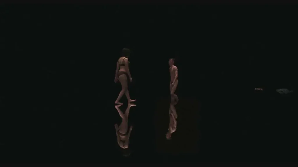La pièce est entièrement plongée dans le noir. Une femme en sous-vêtement s'approche d'un homme entièrement nue sui semble s'enfoncer dans un eau noire.