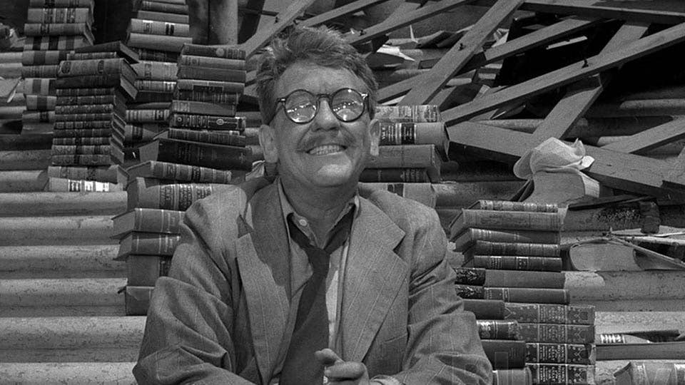 Un homme à lunette devant une pile de livres dans ce qui semble être un univers apocalyptique. The Twilight Zone.
