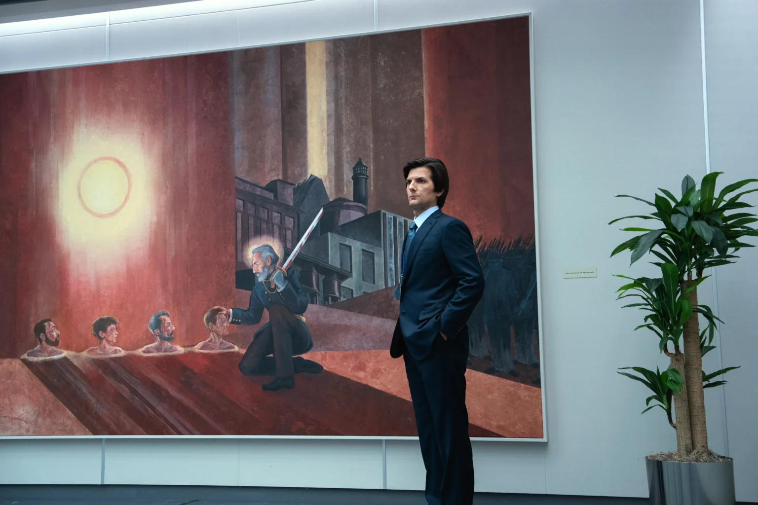 Mark Scout, de Severance, se tenant droit juste à côté d'une plante et un tableau relevant du culte de Kier.