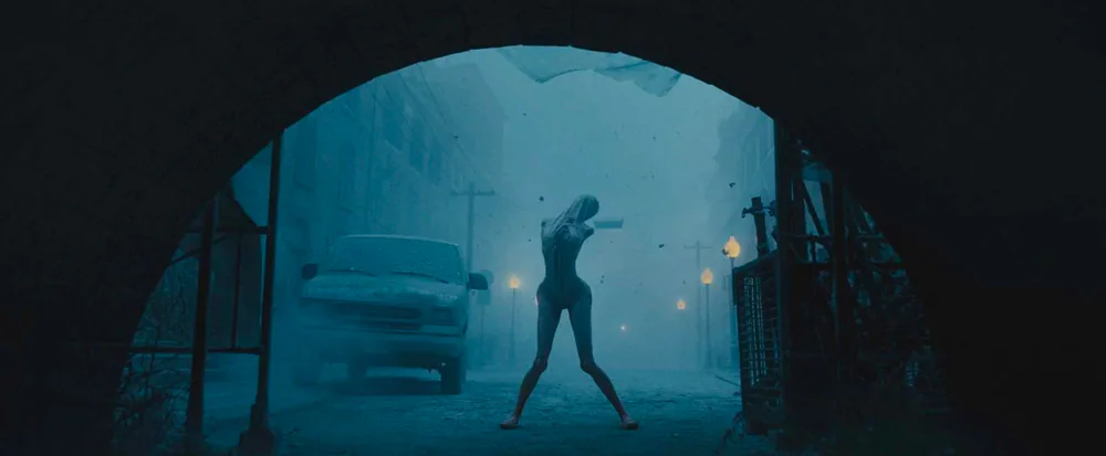 Un femme, au corps étrange, comme si elle était sous camisole mais celle(ci faisait pasrtie de sa peau, approche d enous en passant sous un pont. L'image est d'un bleu inquiétant. Tirée du film Retour à Silent Hill (2026).
