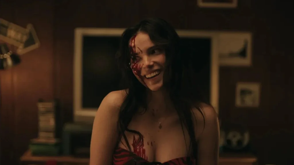 Une femme, le visage ensanglanté souri bizarrement, elle ne semble pas dans on état normal. L'image est tirée du film Obsession (2026)