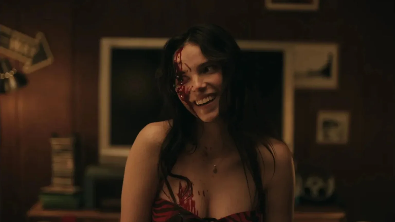 Une femme, le visage ensanglanté souri bizarrement, elle ne semble pas dans on état normal. L'image est tirée du film Obsession (2026)