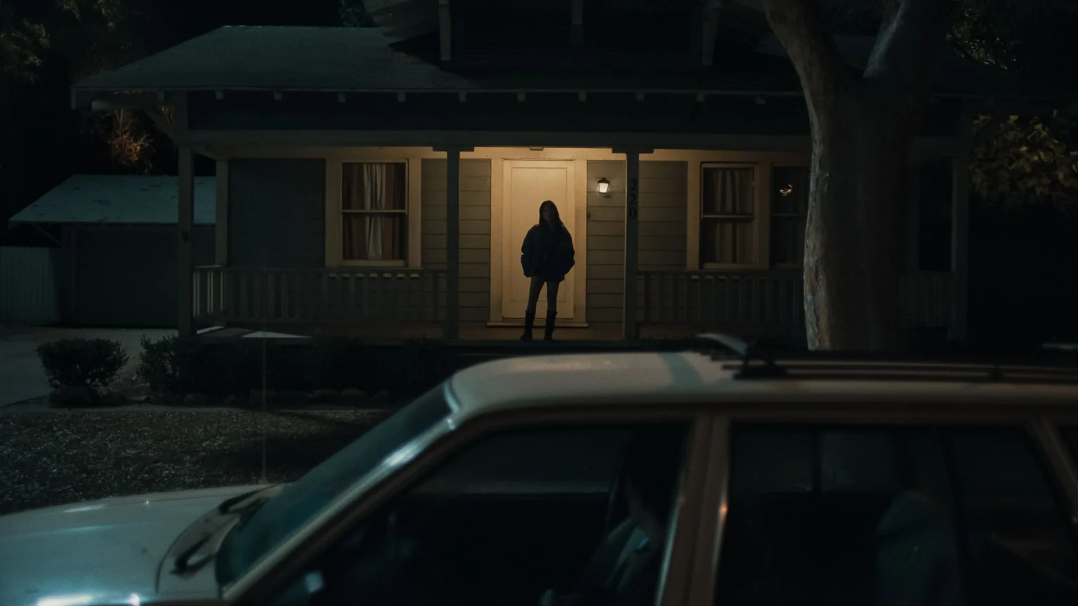 L'image est tirée du film Obsession (2026). On y voit une femme dessous le porche éclairée d'une maison, la nit, regardant droite devant elle vers la voiture au premier plan.