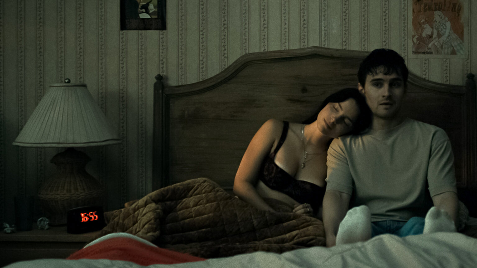Un homme et une femme sont assis dans un lit, le réveil indique 6h55 du matin. La femme pose la tête sur son épaule à lui, mais lui ne semble pas serein. L'image est tirée du film Obsession (2026)