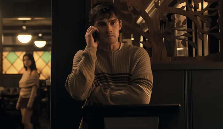 Un homme au premier plan, dans un restaurant, téléphone à quelqu'un et semble inquiet, derrière au second pan, une femme le regard fixement. L'image est tirée du film Obsession (2026)