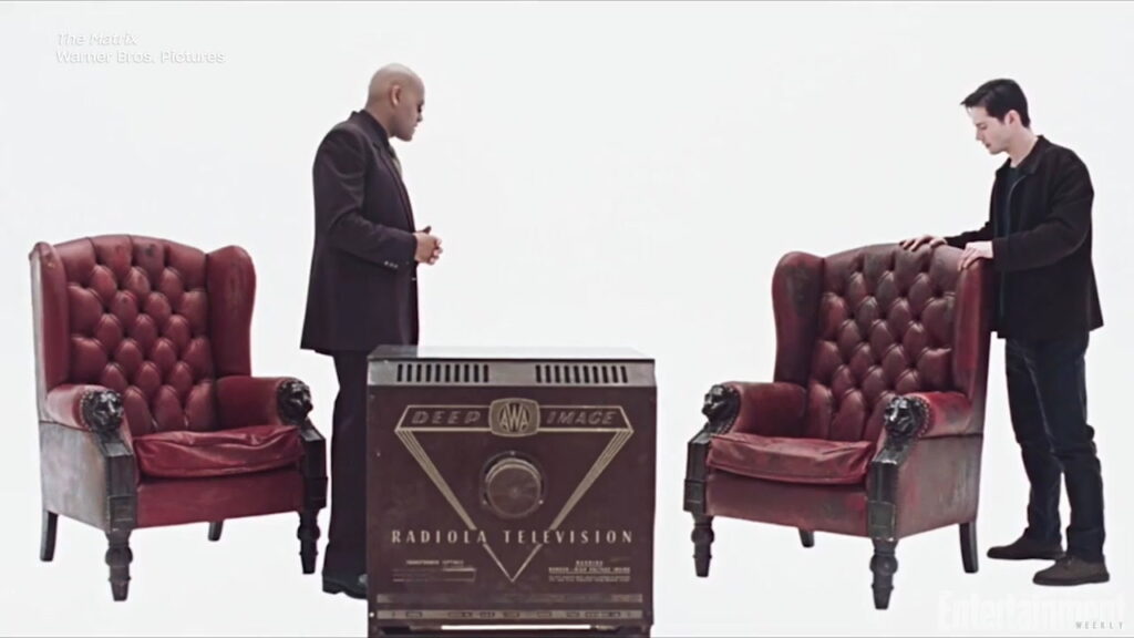 Deux hommes discute, prêts à s'asseoir sur des fauteuils rouges en cuir, placés devant une télé que l'on voit que de derrière. Le reste de la pièce est entièrement vide et sans limites. Image tirées de Matrix